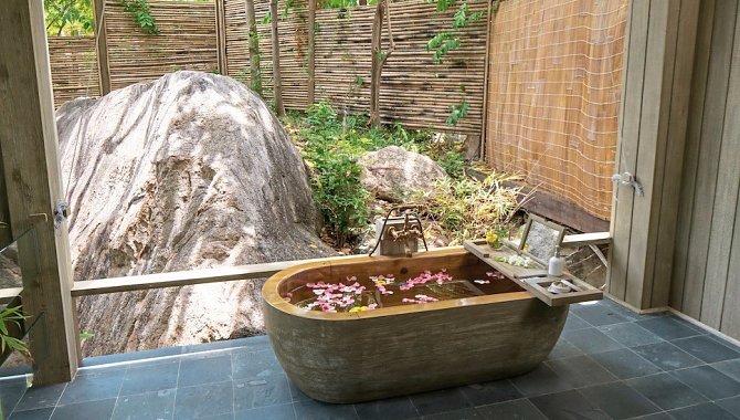 Naturstein-Badewanne mit Blütenblättern im Wasser, draußen vor einer Felswand.