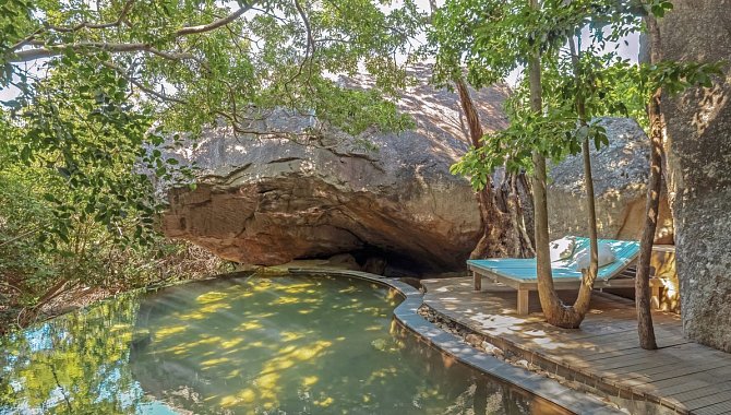 Natürlicher Pool im Schatten großer Felsen und Bäume mit Liegefläche.