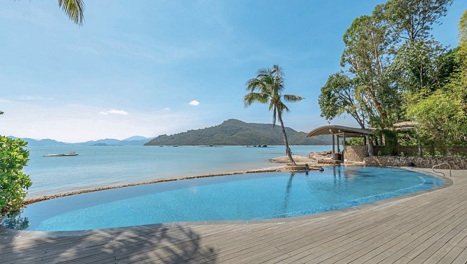 Infinity-Pool mit Holzterrasse direkt am Meer, umgeben von Palmen und Natur.