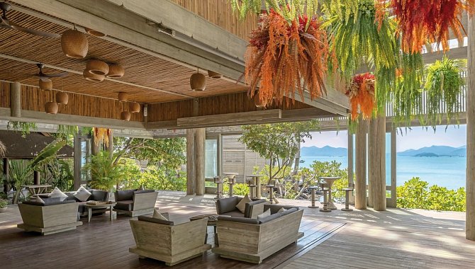 Überdachte Lounge mit Sitzgruppen, Holzdecken und bunten Hängepflanzen, Meerblick im Hintergrund.