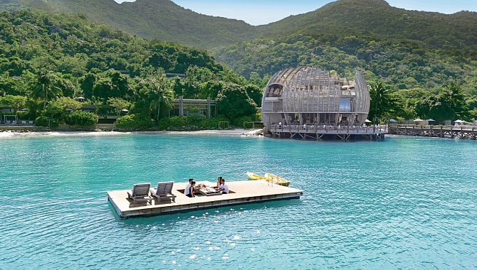 Schwimmende Plattform mit Sonnenliegen und Personen auf dem türkisblauen Meer, im Hintergrund bewaldete Berge und ein modernes Holzgebäude.
