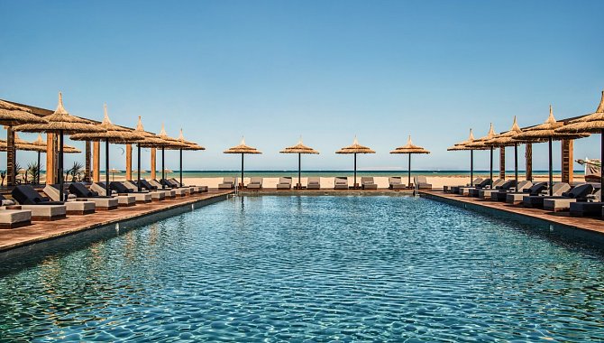 Der große Hotelpool ist gesäumt von Sonnenliegen und Sonnenschirmen aus Stroh. Unter jeden Schirm stehen zwei Liegen.