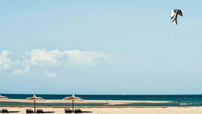 Aussicht auf den Strand. Sonnenliegen und Sonnenschirme stehen Links im Bild, rechts surft ein Kitesurfer auf dem flachen Wasser.