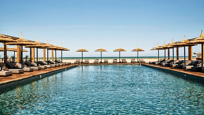 Der große Hotelpool ist gesäumt von Sonnenliegen und Sonnenschirmen aus Stroh. Unter jeden Schirm stehen zwei Liegen.