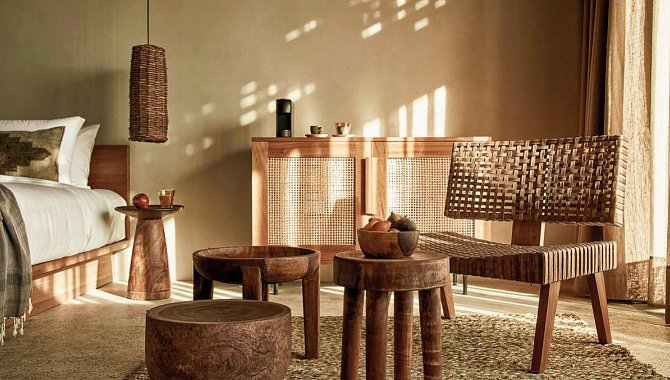 Sitzecke in einer der Zimmer. Ein brauner runder Beistelltisch mit einer Obstschale darauf, Ein Holzhocker und ein gewebter Sessel in Braun. Dahinter eine Hölzerne kommode. Von links fällt licht in das Zimmer, alles ist in natürlichen Erdtönen gehalten.