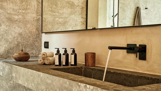 Das Waschbecken im Badezimmer. Ein großer durchgehender Spiegel hängt an der Wand. Die Waschtheke ist aus Steinmaterialien und das große Waschbecken ist in diese eingelassen, der Stilvolle mattschwarze Wasserhahn kommt direkt aus der Wand.