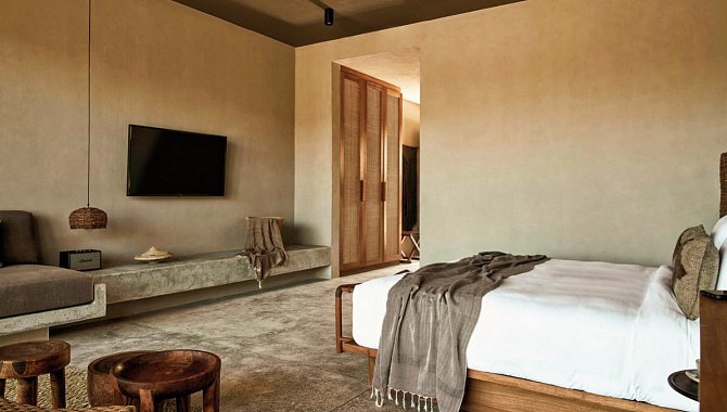 Das Bett des Zimmers steht in der Linken Ecke des Raumes, es ist aus Holz und mit weißen Laken bezogen. Gegenüber des Bettes hängt der Fernseher, darunter das Sideboard, welches an der Wand befestigt ist und aus Stein gemacht ist, das Sofa grenzt direkt daran an, sodass der Eindruck entsteht beides sei aus einem Guss.