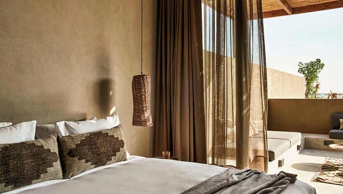 Eine Zimmerecke einer Junior Suite. Durch die geöffnete Schiebetür kann man die Veranda sehen. Man sieht einen Teil des großen weiß bezogenen Doppelbettes und die Hängelampe in der Ecke. Es gibt durchsichtige vorhänge für den Tag und dicke Braune zum Abdunkeln des Raumes.