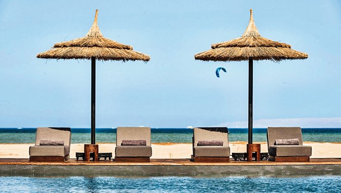 Der Pool mit zwei Snnenschirmen. Unter den Schirmen stehen jeweils zwei Poolliegen. Im Hintergrund sieht man den Sandstrand und das Meer.