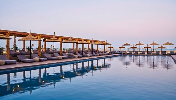 Der große rechteckige Hotelpool gesäumt von Poolliegen und Sonnenschirmen. Wolkenloser Himmel an einem frühen Abend.