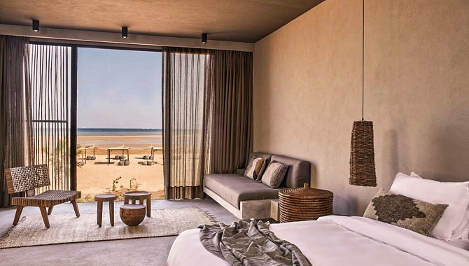 rechts im Vordergrund sieht man einen Teil des Bettes, dahinter das Sofa mit einem Teppich und Kaffeetischen. Das Sofa steht neben der großen Fensterfront mit einer großen geöffneten Schiebetür.