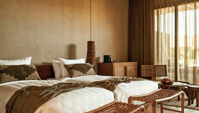 Zwei große Betten stehen nebeneinander. Am Fenster steht ein gewebter Sessel mit großem Runden beistelltisch. In der Ecke steht eine hölzerne Kommode. Vor den Betten