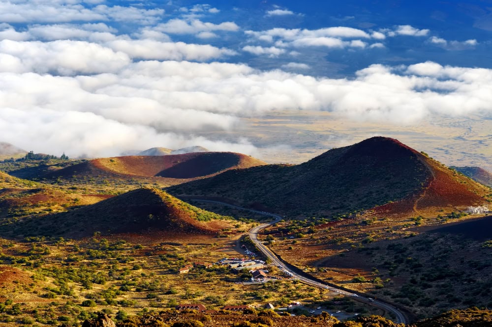 Vulkanlandschaft des Mauna Loa auf Hawaii mit roten Kraterhügeln und Wolkenmeer im Hintergrund.
