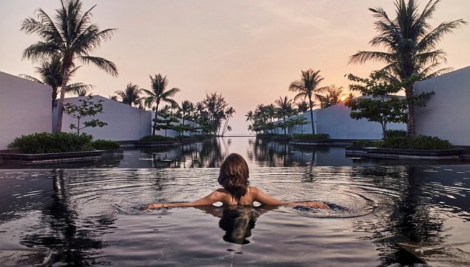 Frau schwimmt im Infinity-Pool bei Sonnenuntergang mit Palmen im Hintergrund